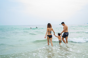 Fototapeta premium Happy asian family - father, mother, baby daughter hold hands and run with fun along edge of sea on sand beach. Active parents and people outdoor activity on tropical summer vacations with children.