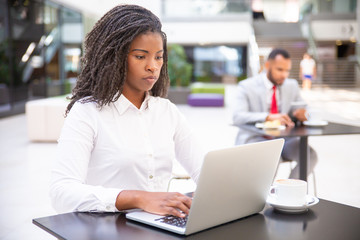 Confident young businesswoman using computer in co-working space. African American business woman looking at laptop screen, man using tablet in background. Young businesswoman concept