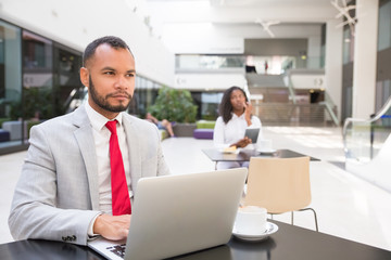 Pensive business professional thinking over ideas for project. Business man sitting at table in office lobby, using laptop, young woman using tablet in background. Working in public space concept