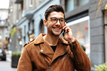 City style. Technology. Handsome young man in casual clothes and glasses is talking on the mobile phone and smiling while walking in the city