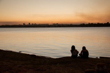 Naklejka na ściany i meble Two Ladies Sitting on the Shore of Lake Paranoa watching the sunset over the city of Brasilia, Brazil