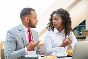Excited diverse professionals discussing project during breakfast. Business man and woman sitting at laptop in cafe, drinking coffee, eating, talking. Lunch break concept