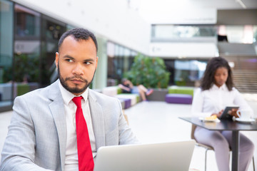 Serious business leader working on computer while waiting someone in office hall. Young woman using gadget in background. Workspace concept