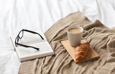 Fototapeta na wymiar comfort, hygge and interior concept - coffee, croissant, blanket and book on bed at cozy home