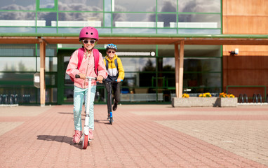 education, childhood and people concept - happy school children in helmets with backpacks riding scooters outdoors