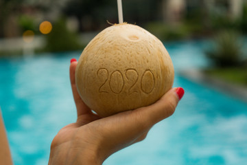 Coconut with straw with 2020 drawing on the beach near the sea with New year winter holiday concept. Red haird girl holding coconut near pool