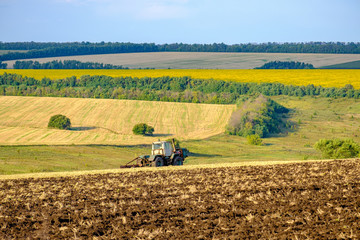 An agricultural tractor plows a field with a plow after harvested wheat. In the background are fields of sunflowers.
