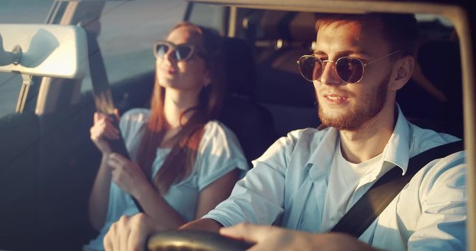 Happy young couple in sunglasses and casual outwear driving in car on sunset, enjoying travelling adventures