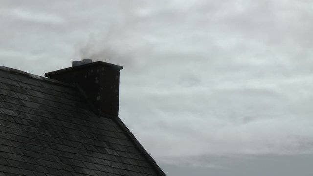 Close up of an old fashioned chimney at the top of a farmhouse framed against an overcast sky, with a thin waft of smoke drifting from it