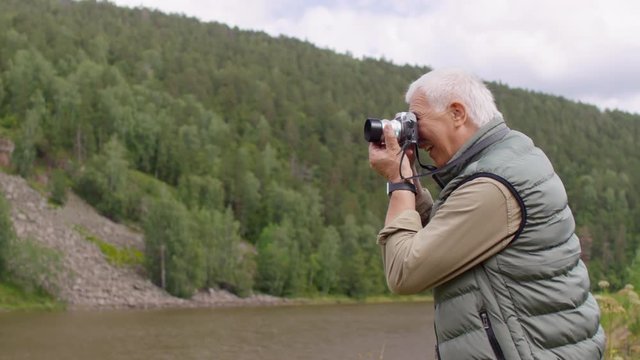 Tracking shot of happy elderly man with grey hair and mustache standing by river and taking picture on digital camera during hike on chilly summer day