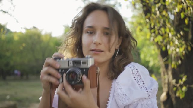 Portrait of attractive young photographer with curly hair looking at the camera while taking photo using old camera in the garden or park. Rural life. Retro style