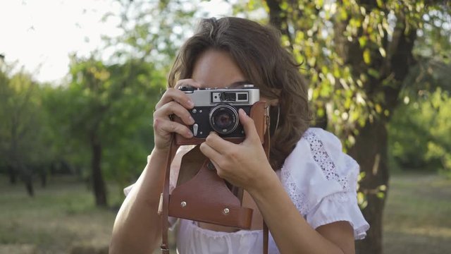 Portrait of cute young photographer with curly hair looking at the camera while taking photo using old camera in the garden or park. Rural life. Retro style