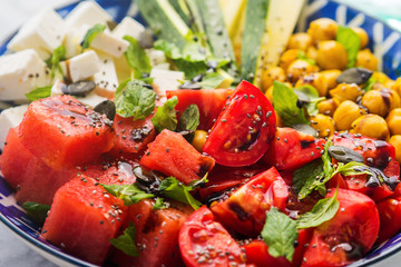 Colorful turmeric chickpea watermelon salad with feta cheese, tomatoes, zucchini, mint and balsamic vinegar on marble table. Summer fresh easy lunch