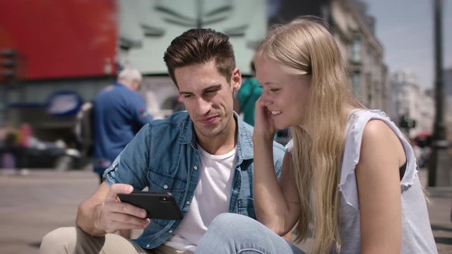 Happy young couple sitting in Piccadilly Circus in London. Beautiful young couple dating smiling and watching messages and pictures on a smartphone in UK. Blond woman, handsome man.
