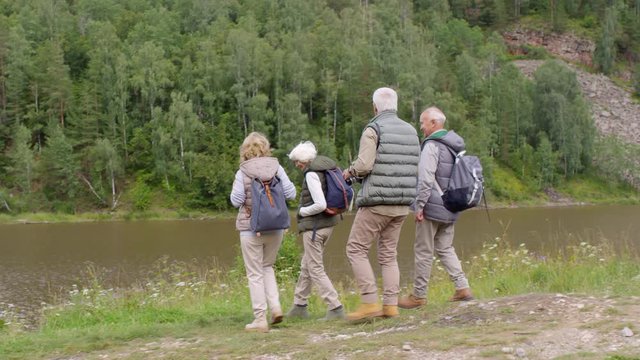 Tracking shot of group of senior male and female tourists with backpacks hiking on chilly summer day, then stopping by river and enjoying view