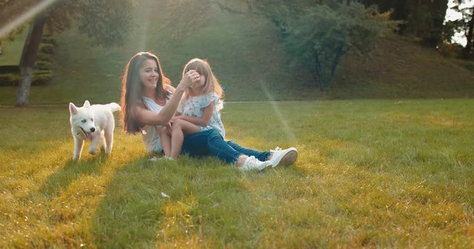 Caucasian cheerful pretty mother and small cute daughter sitting on the green grass in the sunny park with a while haski dog.