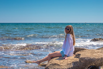 blonde with long hair sits on stones on the shore of the blue sea and stretched her bare legs forward