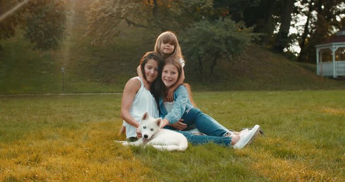 Portrait shot of the beautiful young Caucasian happy mother with two cute pretty small daughters and haski dog sitting on the green grass and smiling to the camera.