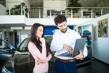 Salesman with laptop and client standing near new car indoors