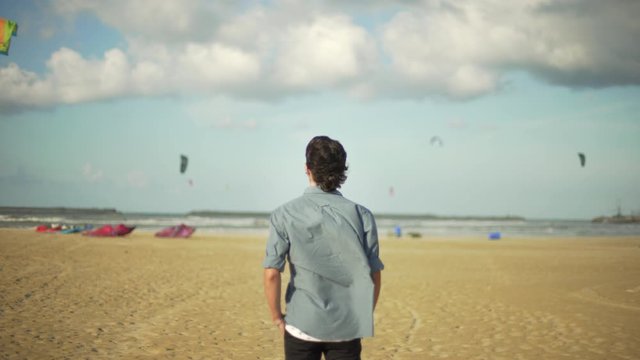 Young sensitive guy looking at kitesurfes flying in the sky under a blue and cloudy sky by the sea. Beach in a windy day.