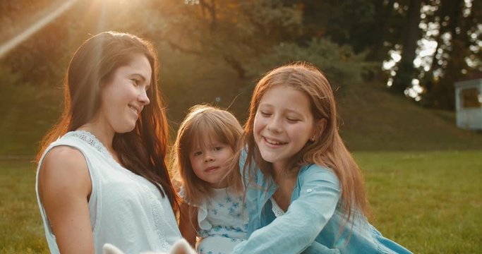 Close up of the joyful Caucasian prety mother and two small girls her daughters having fun and laughing to the camera in the park on sunny day in summer.