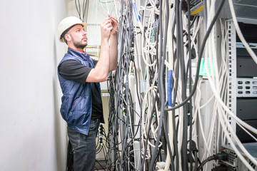 An engineer in a white helmet connects the telecommunication wires in the rack of the server room. Chaotic plexus electrical wires. A technician works with many wires in a data center.