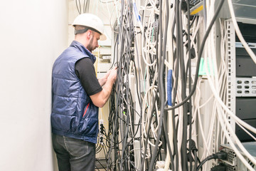 A technician works with many wires in a data center. An engineer in a white helmet connects the telecommunication wires in the rack of the server room. View from the back