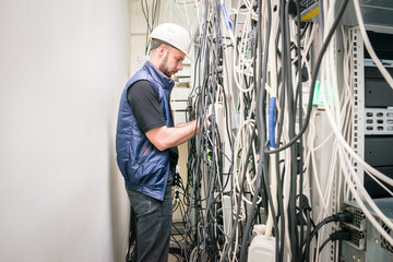 A technician working connects a lot of cables in a server rack. Chaotic plexus electrical wires. An engineer in a white helmet unravels many wires in a data center.