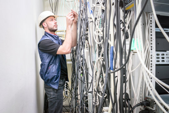 An engineer in a white helmet connects the telecommunication wires in the rack of the server room. Chaotic plexus electrical wires. A technician works with many wires in a data center.