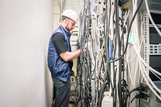 A technician working connects a lot of cables in a server rack. Chaotic plexus electrical wires. An engineer in a white helmet unravels many wires in a data center.