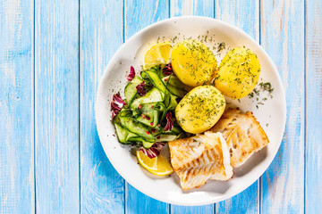 Fried fish with potatoes and vegetable salad on white plate on blue planks