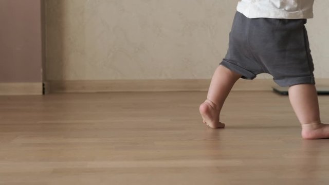 Closeup of baby tiny barefoot legs doing first steps and walking on floor at home without any help. Slow motion