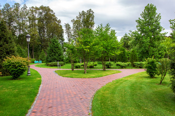 Footpath in perspective a park with green lawns and trees, among which are mounted iron street lantern on a summer day with clouds nobody.