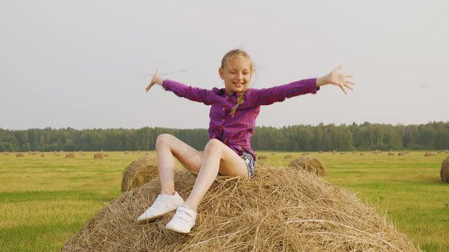 Girl scattering straw on field. Adorable happy girl in checkered shirt and denim shorts sitting on haystack and smiling at camera. Harvest concept