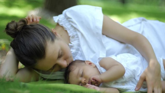 Mother kisses her Baby sleeping on a green grass Outdoors. Happy smiling young Mother and Child in Green Summer Park. Beautiful family in spring park enjoying nature.