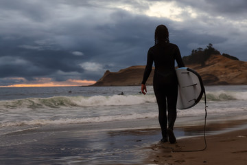 Adventurous Girl with a Surf Board is going surfing in the Ocean during a cloudy summer sunset. Taken in Pacific City, Oregon Coast, United States of America.