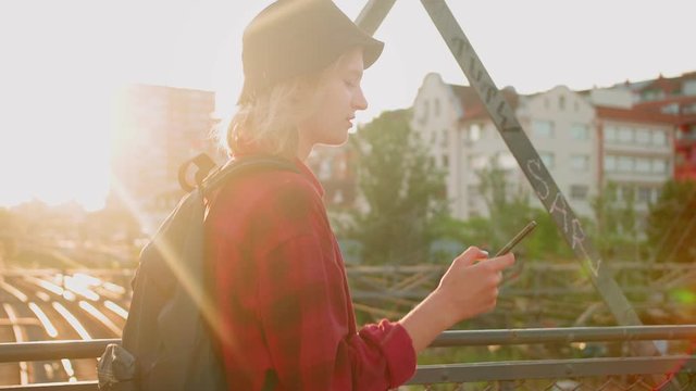 Caucasian teenage girl in hat walking at pedestrian bridge using black smartphone, early morning sunrise pleasant millennial holding mobile device to be online and up to date with best friends