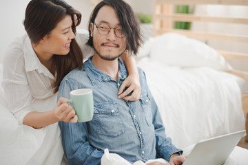 Young Asian man sit on the ground and working with laptop computer which woman lie on the bed behind man and they are smiling feeling happy in the bedroom at home. the life at home.Couple love.