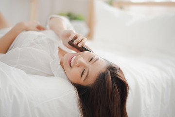 Obraz na płótnie Canvas Young Asian woman lie supine which talking and using a smart phone on the white bed at home looking felling happy. the life at home concept,