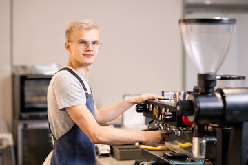 Successful barista in uniform using coffee machine while standing by workplace