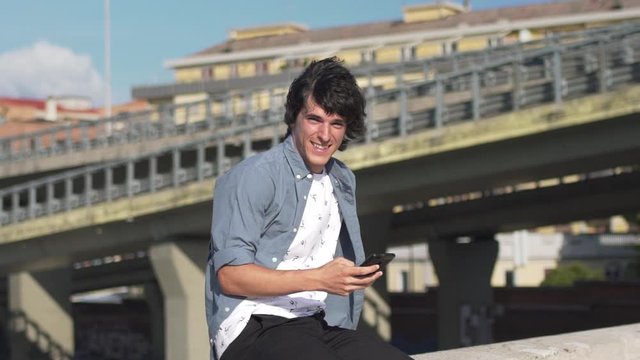 Handsome guy using cellphone or smartphone and smiling in the city near a bridge or overpass. Attractive young man using mobile and looking at camera in slow motion.