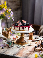 homemade tasty whole cheesecake decorated with figs, blackberries, blueberries and purple sauce on top served on wooden cake stand on grey table with flowers and berries, selective focus