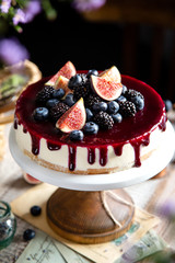 homemade tasty whole cheesecake decorated with figs, blackberries, blueberries and purple sauce on top served on wooden cake stand on grey table with flowers and berries, selective focus