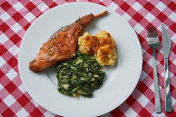 Flat lay picture of roasted rabbit leg, marinated in garlic and rosemary served with spinach, chopped onion and czech potato dumplings with fork and knife. Traditional dish cuisine of Czech republic.