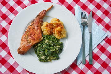 Flat lay picture of roasted rabbit leg, marinated in garlic and rosemary served with spinach, chopped onion and potato dumplings with fork, knife and napkin.Ttraditional dish cuisine of Czech republic