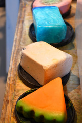 An image of four various decorative soap bars of different colors (pink, blue, white, orange and green) and shapes (round, rectangular and circular segment) made of natural ingredients