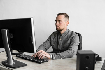 Young businessman or student in a shirt sitting against monitor of computer. Working on a pc at a table in the office with a thoughtful expression. Young businessman working on his laptop in office.