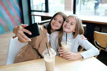Young cheerful female and her daughter in casualwear making selfie in cafe