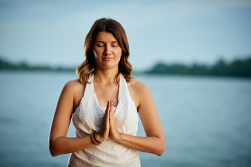 Fototapeta na wymiar Beautiful Caucasian brunette dressed in white standing on dock and standing in mountain yoga position. In background is river.