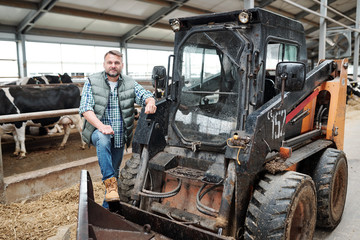 Worker of farmhouse standing by tractor while cleaning aisle of animal farm